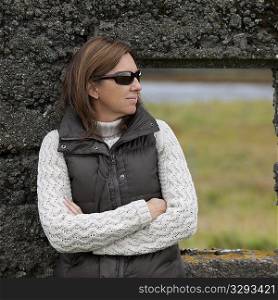 Casual portrait woman in sunglasses, down vest, and knit sweater , before rugged stone window frame