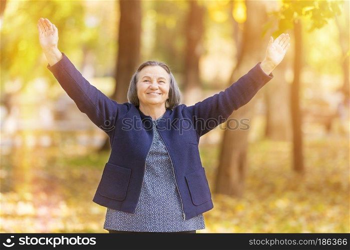 Casual senior woman with arms outstretched standing in autumn park. 