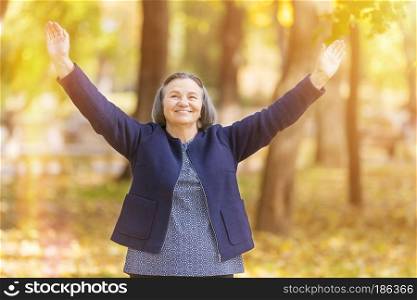 Casual senior woman with arms outstretched standing in autumn park. 