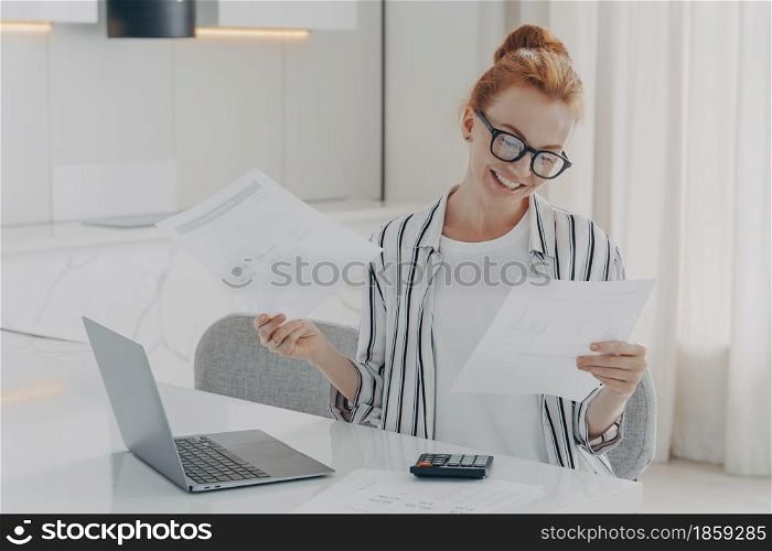 Casually dressed excited redhead woman sitting at table at home, feeling happy after checking domestic bills, holding holding documents with bank notification and smiling, using laptop and calculator. Young excited redhead woman sitting at table at home, feeling happy after checking domestic bills,