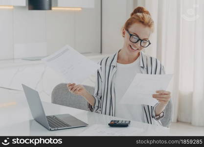 Casually dressed excited redhead woman sitting at table at home, feeling happy after checking domestic bills, holding holding documents with bank notification and smiling, using laptop and calculator. Young excited redhead woman sitting at table at home, feeling happy after checking domestic bills,