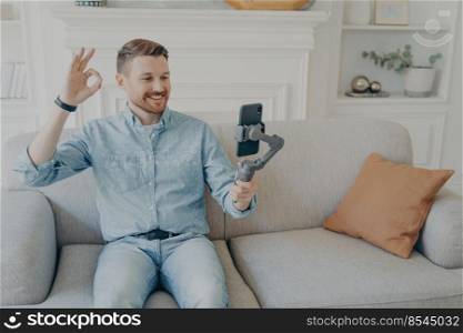 Casually dressed young man showing thumbs up while video chatting using phone attached to gimbal, sitting comfortably on beige couch in living room, having conversationg with family. Young man showing thumbs up while video chatting
