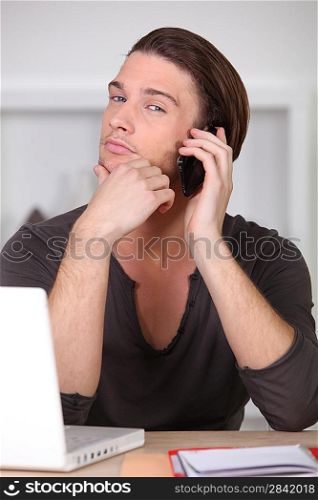 Casually dressed young man using a cellphone at his laptop