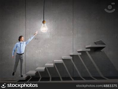 Catch bright idea. Young businessman on ladder reaching hand to touch glowing light bulb