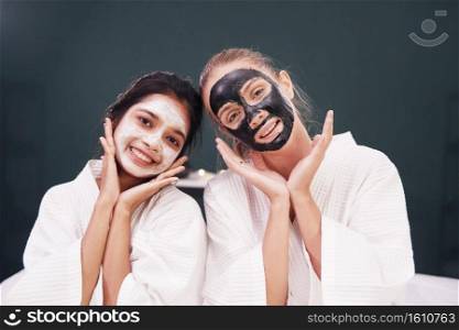 Caucasian and Asian woman in white bathrobe and black facial mask on face for treatment skin care together at spa. Lifestyle and  relationship lesbian lover. Beauty salon concept.