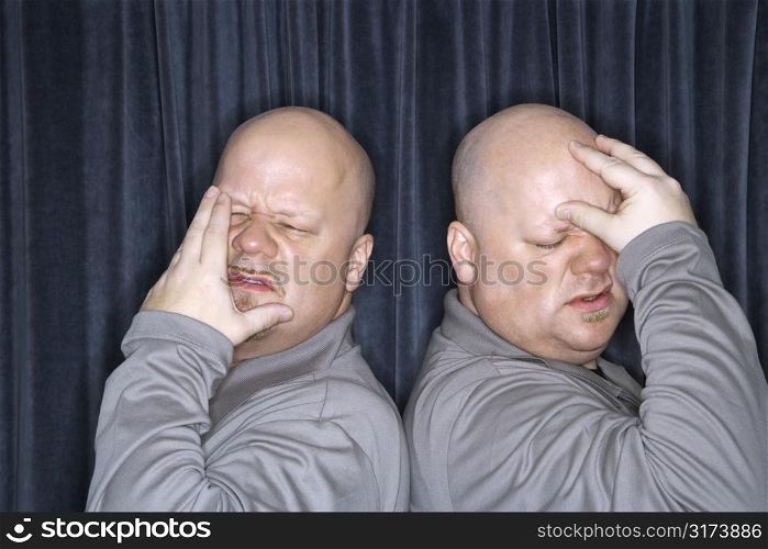 Caucasian bald identical twin men standing back to back and grimicing with hands to head.