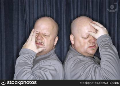 Caucasian bald identical twin men standing back to back and grimicing with hands to head.
