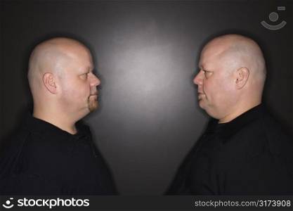 Caucasian bald mid adult identical twin men standing face to face staring.