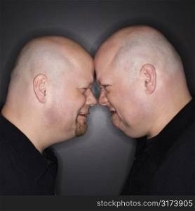 Caucasian bald mid adult identical twin men standing face to face with angry expression.