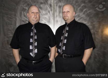 Caucasian bald mid adult identical twin standing together looking at viewer.