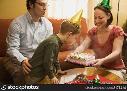 Caucasian boy in party hat blowing out candles on birthday cake with family watching.