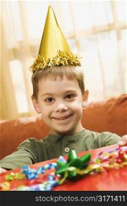 Caucasian boy wearing party hat holding gift and smiling at viewer.