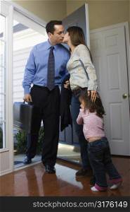 Caucasian businessman at open door kissing wife while daughter hugs her leg.