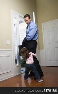 Caucasian businessman at open door with briefcase with daughter tugging on his leg.
