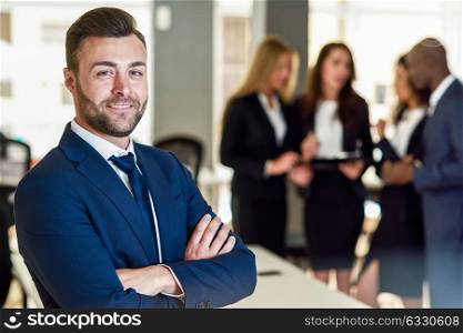 Caucasian businessman leader looking at camera in modern office with multi-ethnic businesspeople working at the background. Teamwork concept. Young man with beard wearing blue suit.
