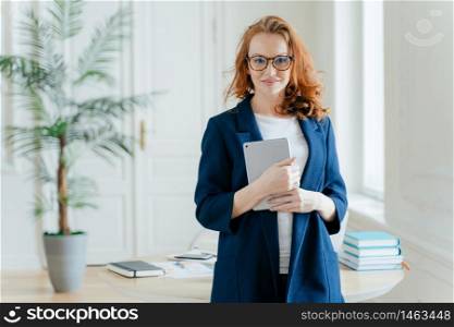 Caucasian businesswoman holds tablet computer, checks balance account, wears optical glasses and formal outfit, waits for notification stands indoor, desktop with book in background. Technology