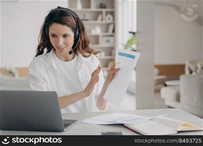 Caucasian businesswoman in headset is reporting online from home. Corporate internet session on laptop. Young pretty girl shows charts and talking in front of camera. Remote business on quarantine.. Caucasian businesswoman in headset is reporting online. Corporate internet session on laptop.