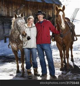 Caucasian couple holding horses in winter with stable in background.