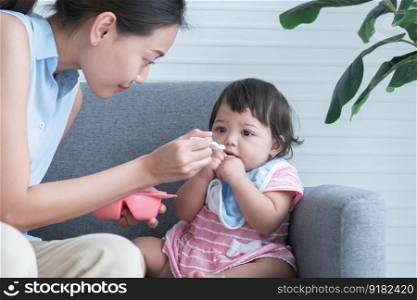 Caucasian Cute 7 month newborn baby girl deny eating blend mashed food, young Asian mother holding spoon feeding child, sitting on sofa with apron at home, baby is full and mouth mess up with food