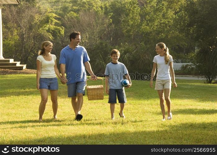 Caucasian family of four walking in park carrying picnic basket.