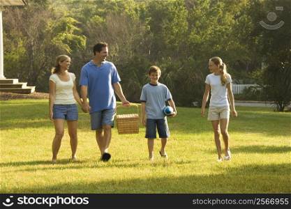 Caucasian family of four walking in park carrying picnic basket.