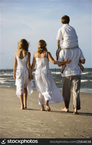 Caucasian family of four walking on beach with dad carrying son on shoulders.