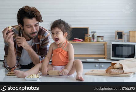 Caucasian family young father and little daughter cooking in the kitchen at home. Cute girl laughing and have fun with rolling pin in hands while dad looking at his baby and holding an ice cream cookie