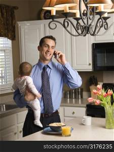 Caucasian father in suit holding baby and talking on cellphone in kitchen.