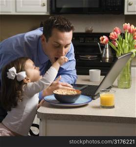 Caucasian father in suit using laptop computer with daughter feeding him breakfast in kitchen.