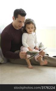 Caucasian father reading book to girl on lap sitting on floor.