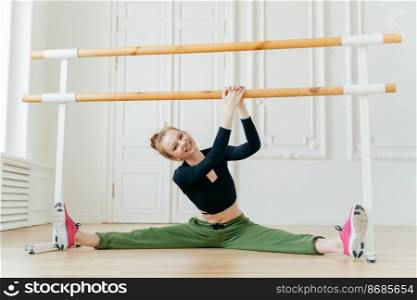 Caucasian female ballerina spreads legs sideways, shows splits, holds hands on barres, has ballet class, wears sportswear and sneakers, has perfect figure, tilts head on right, stretches herself.