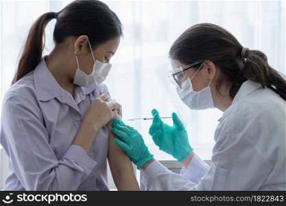 Caucasian female doctor wear mask, face shield and gloves is giving injections or vaccines to upper arm of young Asian woman at hospital. Preventing spread of COVID-19 by vaccinating people concept