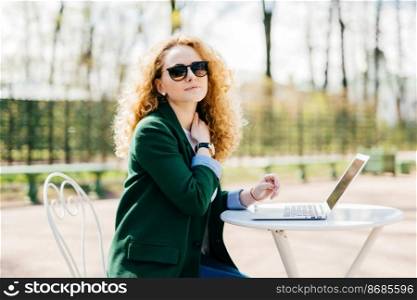 Caucasian female freelancer with fluffy blonde hair wearing trendy clothes and sunglasses working on modern laptop pc holding hand on her neck being little tired and exhausted isolated over nature