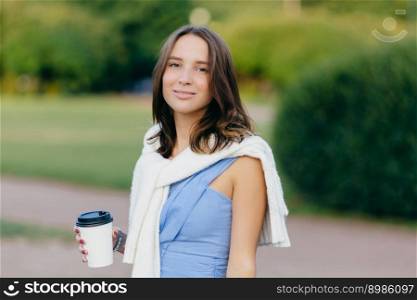 Caucasian female model with pleasant appearance, dressed in casual clothes with sweater on shoulders, holds disposable cup of coffee, breathes fresh air, stands against green nature background