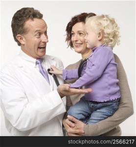 Caucasian female toddler holding stethoscope up to middle-aged male doctor smiling.