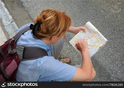Caucasian female tourist wearing a light blue shirt with a backpack is sitting on the edge stone studying city map.