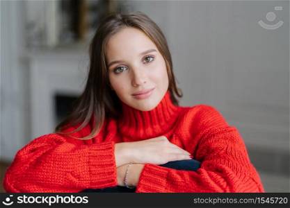 Caucasian female with make up, dark hair, dressd in red warm swater, looks directly at camera with calm expression, poses indoor against blurred background. Relaxed girlfriend enjoys spare time