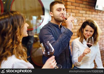 Caucasian friends two sisters or girlfriends and boyfriend with friend standing by the table at the restaurant or winery holding glasses of red wine drinking celebrating wearing shirts Side view