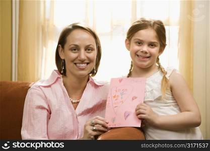Caucasian girl giving mid adult mother a drawing and looking at viewer.