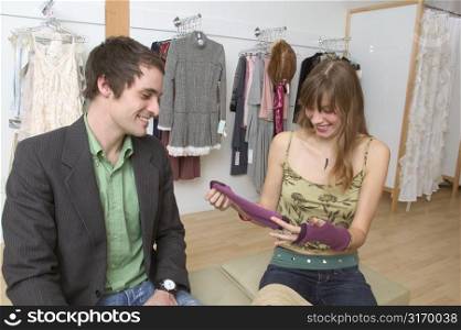 Caucasian Girl Sitting In A Trendy Boutique With Her Boyfriend And Laughing At A Pair Of Gloves