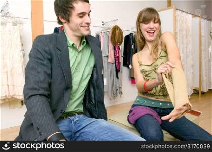 Caucasian Girl Sitting In A Trendy Boutique With Her Boyfriend And Laughing At A Pair Of Gloves