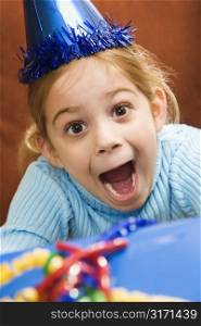 Caucasian girl wearing party hat holding gift and looking at viewer with excited expression.