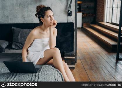 Caucasian girl wrapped in towel after bathing is sitting at laptop on her bed and dreaming. Young woman relaxing and has her time at spa. Weekend morning of freelancer. Bodycare and wellness concept.. Caucasian girl wrapped in towel after bathing is sitting at laptop on her bed and dreaming.