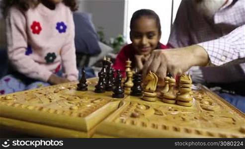 Caucasian grandfather and mixed race granddaughter playing chess at home while cute smiling girl watching the game. Closeup hand of grandpa moving white bishop two fields forward and grandchild capturing it with black pawn. Dolly shot.