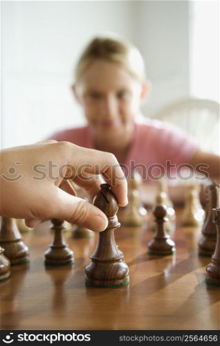 Caucasian hand moving chess piece with pre-teen girl watching in background.