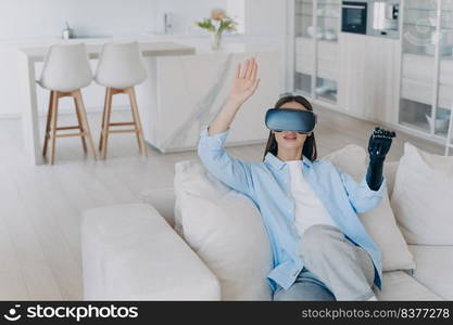 Caucasian handicapped girl in vr glasses. Disabled person gets rehabilitation getting used to sensor cyber arm. Young woman sitting in living room on couch. Amputee with myoelectric robotic limb.. Caucasian handicapped girl in vr glasses. Disabled person gets rehabilitation for sensor cyber arm.