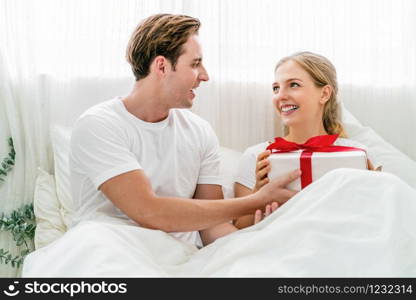 Caucasian Happy couple of lovers is celebrating together on the bedroom in home, Young woman is receiving her boyfriend a gift box for surprise, surprise for valentine or anniversary or christmas.