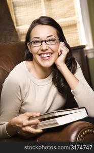 Caucasian/Hispanic young woman sitting in leather chair holding book and smiling at viewer.