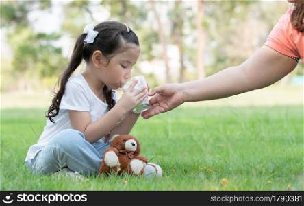 Caucasian little child girl sitting on grass drinking milk from glass hold by her mother at green park on summer. Side view