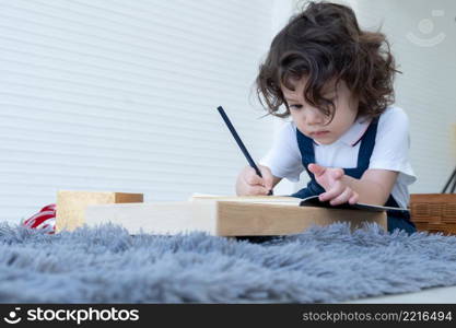 Caucasian little cute kid girl with curly hair sitting and holding pencil writing on book in living room at home with concentrate